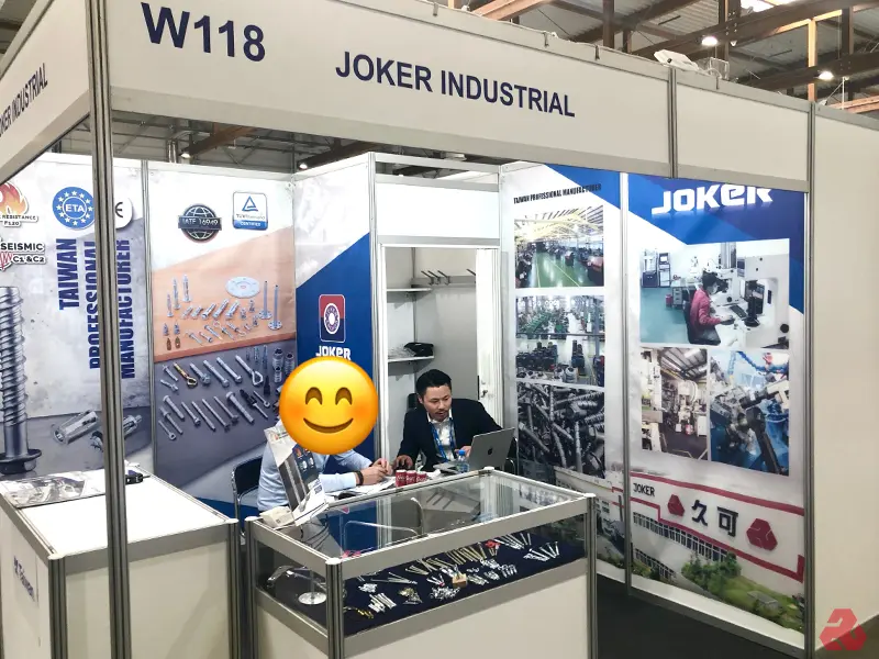 Joker Industrial 在展覽會的 W118 展位，展示台灣製造的螺絲、螺帽及相關產品。展位設計包含公司資訊與產品圖像，一位代表正在使用筆記型電腦，展示櫃中擺放多樣產品。