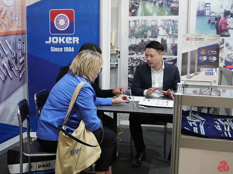 展覽會場景，JOKER ANCHOR 展位，一位穿西裝的亞洲男士正在向兩位客戶展示錨固件產品目錄，桌上擺放螺絲樣品。背景有產品海報和工廠生產線照片，展示錨固件產品及生產實力。