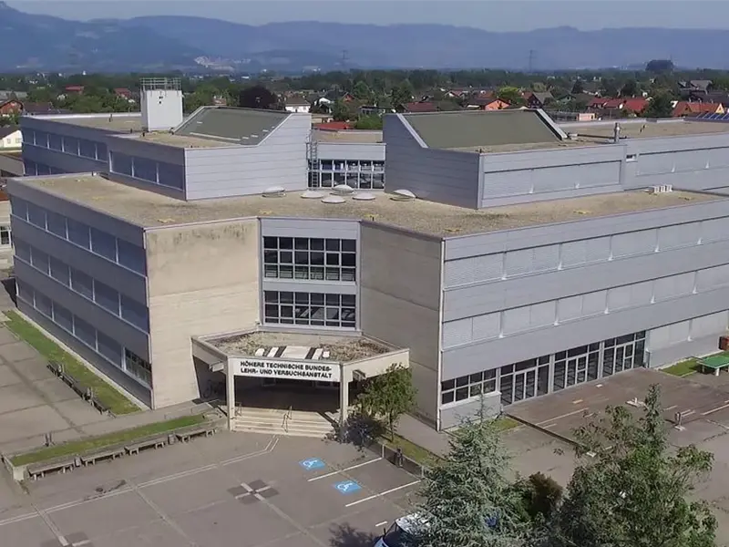 奧地利高等技術聯邦教學及實驗機構，多層灰色建築，設有多個窗戶，屋頂平坦，周圍有綠樹和停車位，背景是連綿的山脈和城鎮建築，呈現一幅寧靜的校園景象。