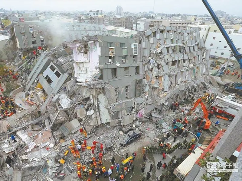 地震災害現場,一棟高樓建築嚴重倒塌損毀,瓦礫堆積如山。大量穿著紅色制服的救援人員正在瓦礫堆中搜尋,橘色挖土機和吊車協助清理,周圍有軍警人員維持秩序。背景為城市景觀。