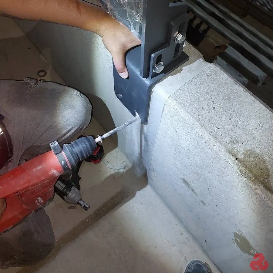施工人員使用紅色電鑽在水泥牆上鑽孔,準備固定灰色的金屬支架。可見支架已初步定位,用螺絲固定部分,鑽孔作業正在進行中,用於完整安裝該支架。