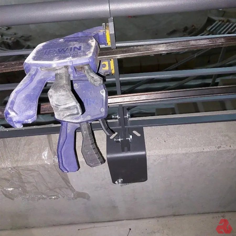 圖片顯示工地現場,一個藍色的IRWIN快速夾具將一塊黑色金屬支架固定在天花板上的金屬框架上。支架透過螺栓連接,用於支撐結構,施工中的細節呈現,顯示裝修工程的進度。